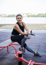 Young brunette woman, wearing black fitness overall, sitting on horizontal bars on sports playground in summer. Fit sportswoman