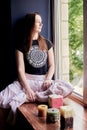 Young brunette woman, wearing black top and light pale pink yoga pants, sitting on windowsill with colorful aroma candles, looking
