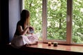 Young brunette woman, wearing black top and light pale pink yoga pants, sitting on windowsill with colorful aroma candles, looking