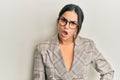 Young brunette woman wearing business jacket and glasses in shock face, looking skeptical and sarcastic, surprised with open mouth Royalty Free Stock Photo