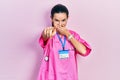 Young brunette woman wearing doctor uniform and stethoscope laughing at you, pointing finger to the camera with hand over mouth, Royalty Free Stock Photo
