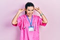 Young brunette woman wearing doctor uniform and stethoscope smiling pulling ears with fingers, funny gesture