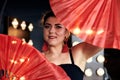 Young brunette woman, wearing little black dress, holding red paper fans in both hands, doing flamenco posing in dark photo studio Royalty Free Stock Photo