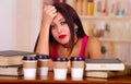 Young brunette woman wearing pink top sitting by desk with stack of books placed on it, holding hand onto head, tired Royalty Free Stock Photo