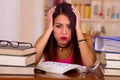 Young brunette woman wearing pink top sitting by desk with stack of books placed on it, holding hands onto head, tired Royalty Free Stock Photo