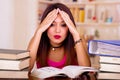 Young brunette woman wearing pink top sitting by desk with stack of books placed on it, holding hands onto head, tired Royalty Free Stock Photo