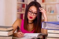 Young brunette woman wearing pink top sitting by desk with stack of books placed on it, resting head onto hand, tired Royalty Free Stock Photo