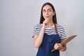 Young brunette woman wearing professional waitress apron and clipboard serious face thinking about question with hand on chin,