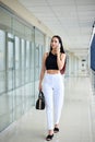 Young brunette woman, wearing white pants and black top, holding black purse, talking on cell phone, standing in light office