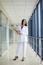 Young brunette woman, wearing white suit, holding black folder with documents,standing in white office building with huge windows.