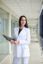 Young brunette woman, wearing white suit, holding black folder with documents,standing in white office building with huge windows.