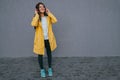 Young brunette woman wearing yellow raincoat and using red headphones listening music during walking outdoor against gray Royalty Free Stock Photo