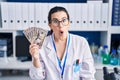 Young brunette woman working at scientist laboratory holding money scared and amazed with open mouth for surprise, disbelief face Royalty Free Stock Photo