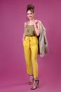 Young woman in yellow pants is holding jacket on her shoulder and laughing. Three quarter length studio shot on pink