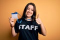 Young brunette worker woman wearing staff t-shirt as uniform showing id card happy with big smile doing ok sign, thumb up with Royalty Free Stock Photo