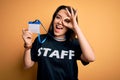 Young brunette worker woman wearing staff t-shirt as uniform showing id card with happy face smiling doing ok sign with hand on Royalty Free Stock Photo