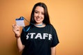 Young brunette worker woman wearing staff t-shirt as uniform showing id card with a happy face standing and smiling with a Royalty Free Stock Photo