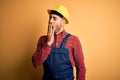 Young builder man wearing construction uniform and safety helmet over yellow isolated background bored yawning tired covering