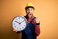 Young builder man wearing safety helmet holding big clock over yellow background screaming proud and celebrating victory and Royalty Free Stock Photo