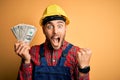 Young builder man wearing safety helmet holding dollars as payment over yellow background screaming proud and celebrating victory Royalty Free Stock Photo
