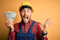 Young builder man wearing safety helmet holding dollars as payment over yellow background very happy and excited, winner Royalty Free Stock Photo