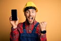 Young builder man wearing safety helmet showing smartphone screen over yellow background screaming proud and celebrating victory Royalty Free Stock Photo