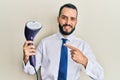 Young business man with beard holding electric steam iron smiling happy pointing with hand and finger Royalty Free Stock Photo