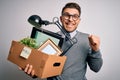 Young business man with blue eyes holding box from the office beeing fired from job screaming proud and celebrating victory and Royalty Free Stock Photo