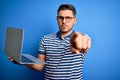 Young business man with blue eyes wearing glasses working using computer laptop pointing with finger to the camera and to you, Royalty Free Stock Photo