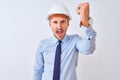 Young business man wearing contractor safety helmet over isolated background angry and mad raising fist frustrated and furious Royalty Free Stock Photo