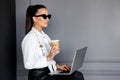 Young business woman in black sunglasses, working on her laptope, holding in hand a cup, posing on grey background. Royalty Free Stock Photo