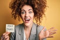 Young business woman with curly hair holding pay taxes to goverment reminder over yellow background very happy and excited, winner