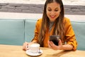 Young business woman drinking cappuccino reading message on smartphone app sitting indoor in urban cafe on break time Royalty Free Stock Photo