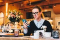 Young business woman in glasses sits in cafe at table, uses smartphone. On table is cup of coffee. Girl working Royalty Free Stock Photo