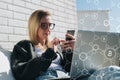 Young business woman in glasses sits at laptop and uses smartphone. In foreground infographics, bitcoin icons. Royalty Free Stock Photo