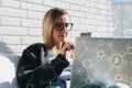 Young business woman in glasses sits at laptop and uses smartphone. In foreground infographics, bitcoin icons. Royalty Free Stock Photo