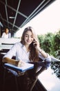 Young business woman sitting at table in cafe, talking oncell phone while taking notes in notebook on table laptop. Student learni Royalty Free Stock Photo