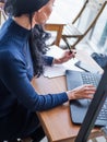 Young business woman in gray dress sitting at table in cafe and writing in notebook. On table is laptop, smartphone and Royalty Free Stock Photo