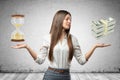 Young business woman holding sand glass and bundles of dollars in her hands on grey wall background Royalty Free Stock Photo