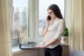 Young business woman with laptop talking on the phone near window Royalty Free Stock Photo