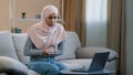 Young business woman muslim islamic arab girl in hijab sitting on sofa working studying online using laptop at home Royalty Free Stock Photo