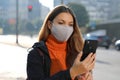 Young business woman with protective mask checking email on mobile phone waiting for a taxi on street in the morning