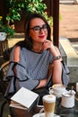 Young business woman sitting in cafe drinking coffee. Beautiful girl in glasses is working against an orange brick wall. View of Royalty Free Stock Photo