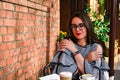 Young business woman sitting in cafe drinking coffee. Beautiful girl in glasses is working against an orange brick wall. View of Royalty Free Stock Photo