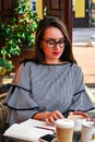 Young business woman sitting in cafe drinking coffee. Beautiful girl in glasses is working against an orange brick wall. View of Royalty Free Stock Photo