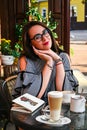 Young business woman sitting in cafe drinking coffee. Beautiful girl in glasses is working against an orange brick wall. View of Royalty Free Stock Photo