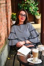 Young business woman sitting in cafe drinking coffee. Beautiful girl in glasses is working against an orange brick wall. View of Royalty Free Stock Photo