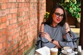 Young business woman sitting in cafe drinking coffee. Beautiful girl in glasses is working against an orange brick wall. View of Royalty Free Stock Photo