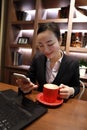 Young business woman sitting in coffee shop at wooden table, drinking coffee and using smartphone.On table is laptop Royalty Free Stock Photo