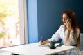 Young business woman sitting at table in cafe, talking on cell phone while taking notes in notebook with cup of coffee Royalty Free Stock Photo